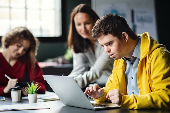 Mehrere Menschen sitzen an einem Tisch. Vor einem Mann in einer gelben Jacke steht ein Computer. 